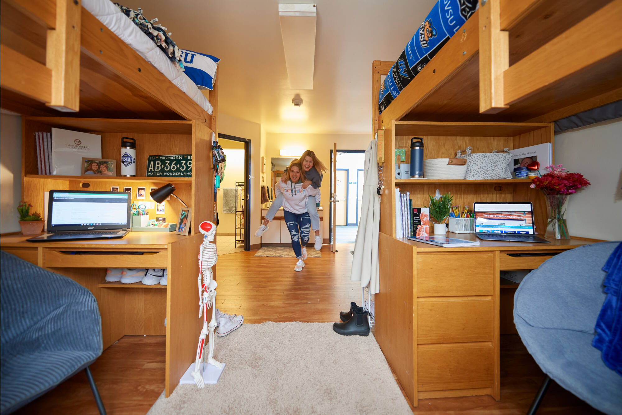Two roommates run and laugh playfully through their room, with two beds and desks, with a bathroom in the background. 